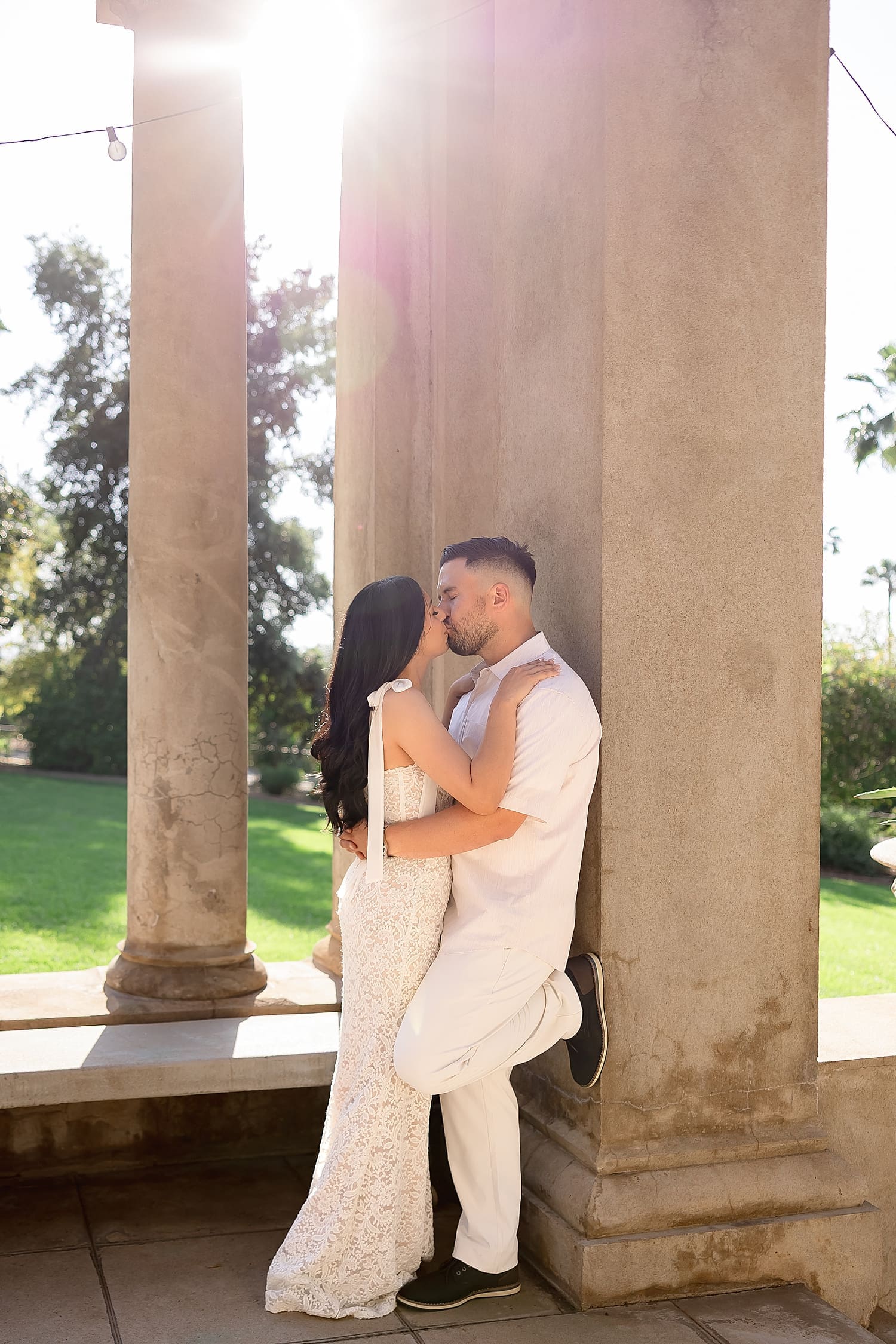 man leans against big stone pillar while bride leans in to kiss him by Desiree Ashley Photography