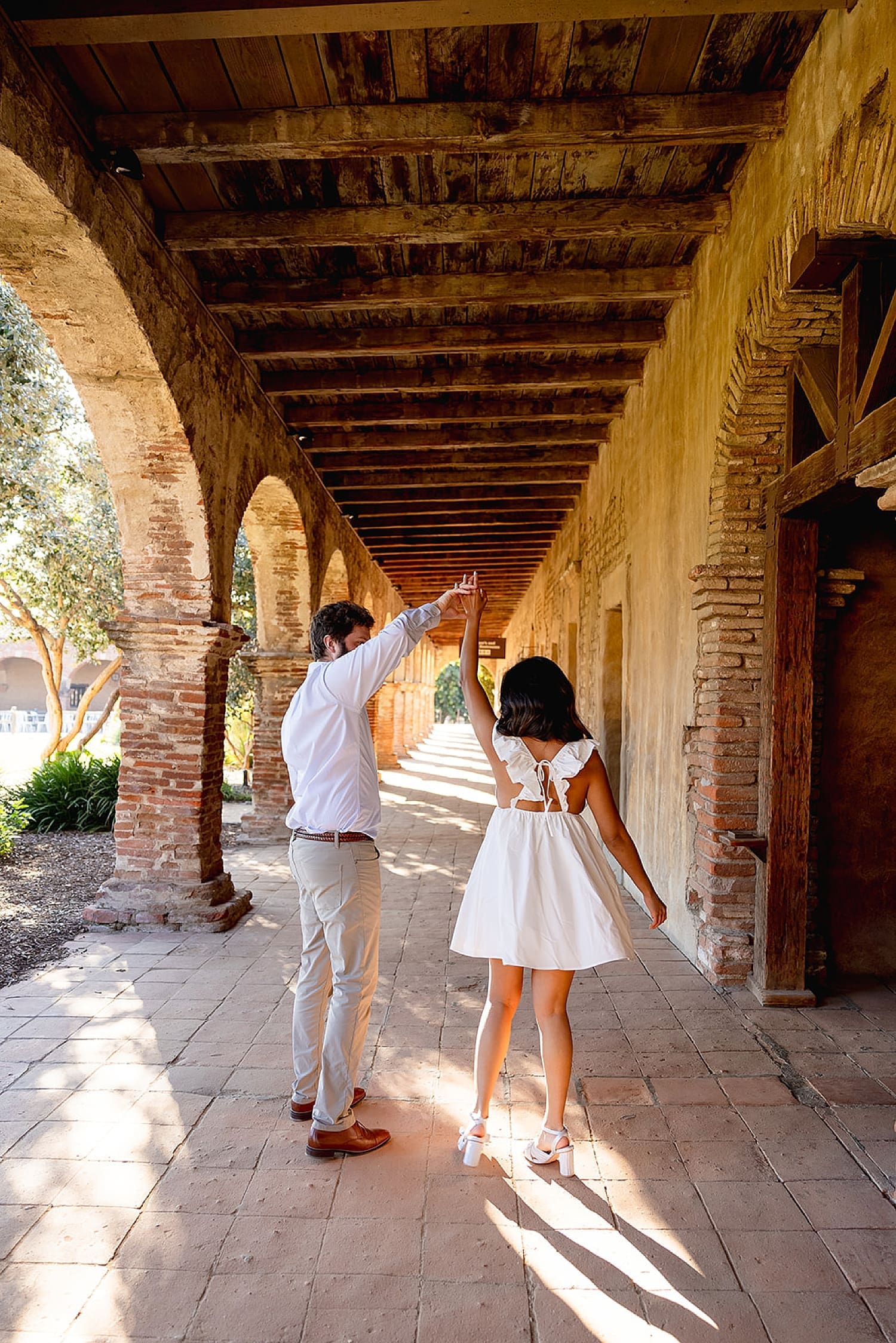 groom-to-be twirls woman in old long outdoor walkway at Mission San Juan Capistrano