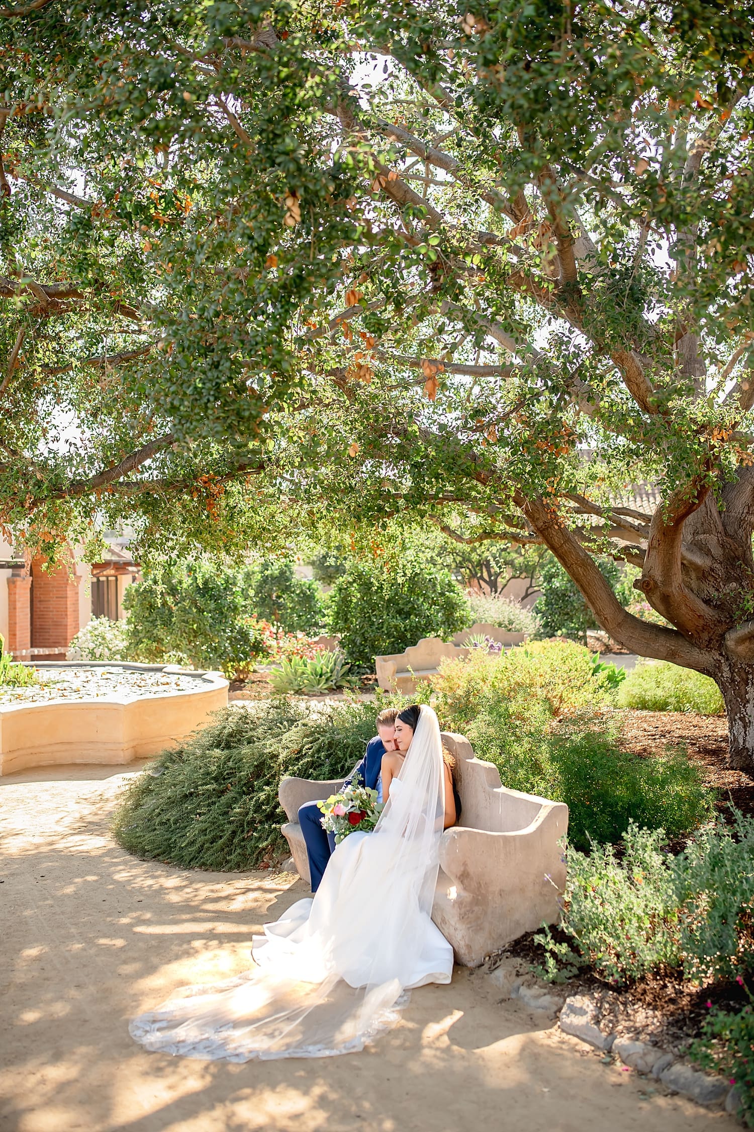bride and groom sit on stone bench in garden at Mission San Juan Capistrano